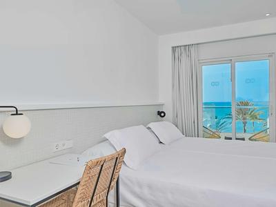 Světý hotelový pokoj se dvěma postelemi, psacím stolem a výhledem na moře přes okno.