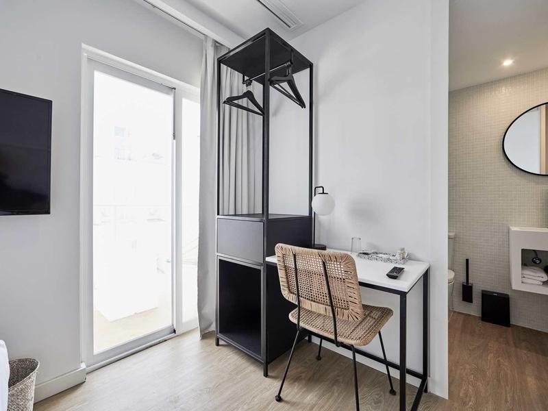 Moderní pracovní prostor se stolem, židlí a otevřeným věšákem v světlém hotelovém pokoji.