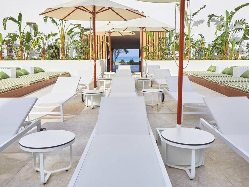 Pohled na moderní venkovní lounge s lehátky, slunečníky a tropickými rostlinami.