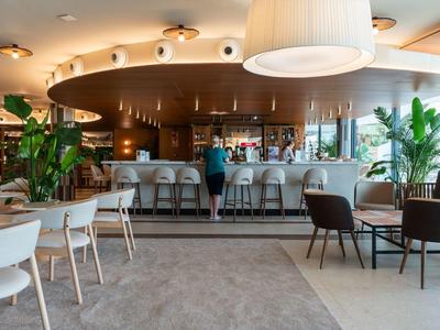 Nowoczesny bar hotelowy z hokerami, roślinami i stolikami w ciepłym oświetleniu.