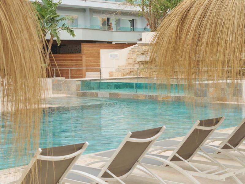 Kilka leżaków przy basenie z daszkami ze słomy przed budynkiem hotelu.