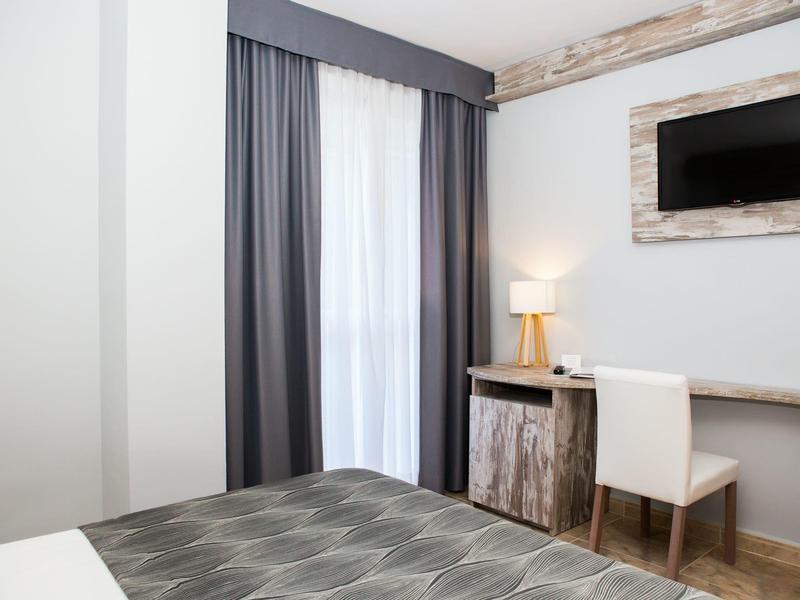 Jasny pokój hotelowy z biurkiem, krzesłem, telewizorem i oknem z zasłonami.