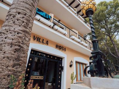 Fassade der Unterkunft „Villa Rosa“ mit Palme, Laterne und Balkon unter blauem Himmel.