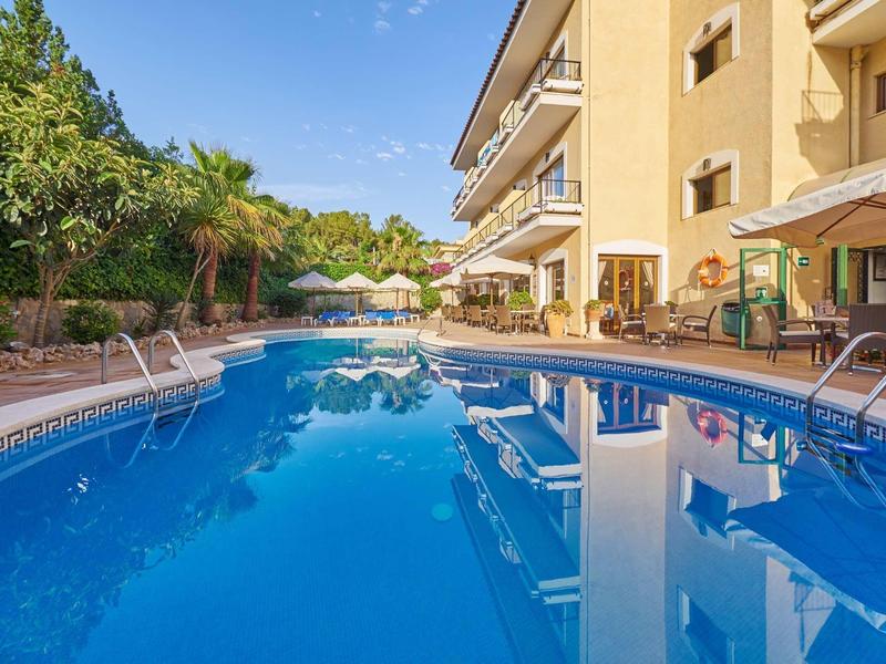 Un giardino di un hotel con piscina, lettini e aree ombreggiate sotto un cielo azzurro.