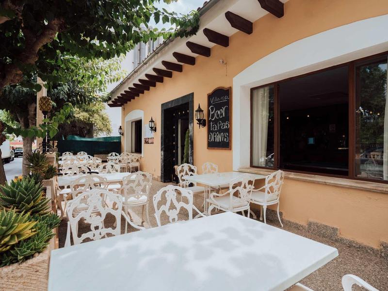 Area esterna dell'hotel con tavoli e sedie bianchi sotto un'area ombreggiata dagli alberi.