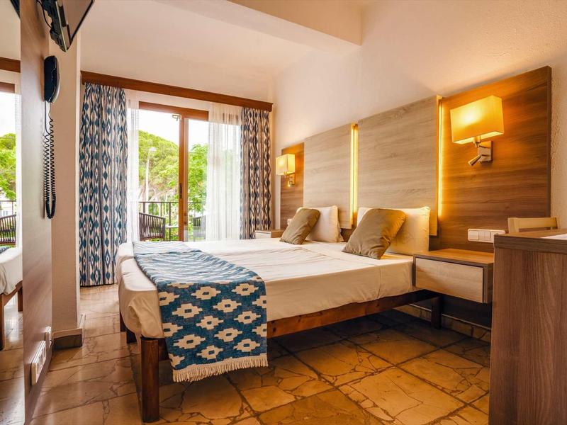 Camera d'hotel luminosa con due letti, mobili in legno e balcone con vista su ambiente verde.