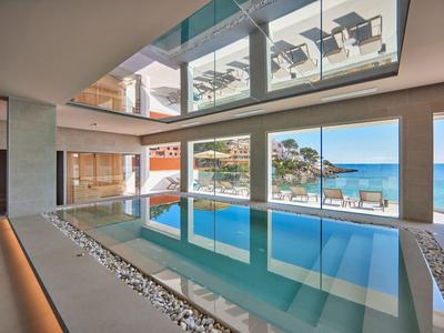 Piscina interior moderna con vista al mar y terraza con sillas.