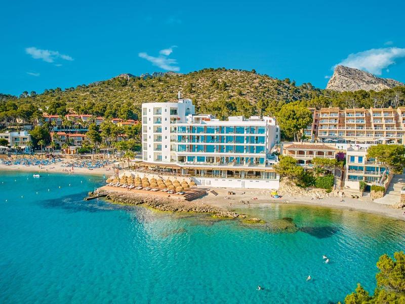 Hotel aan een rustige strand met helder blauw water en beboste heuvels op de achtergrond