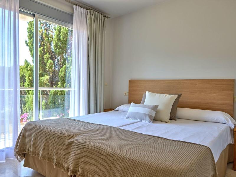 Lichte hotelkamer met groot bed, houten frame en uitzicht op groene bomen door grote ramen.