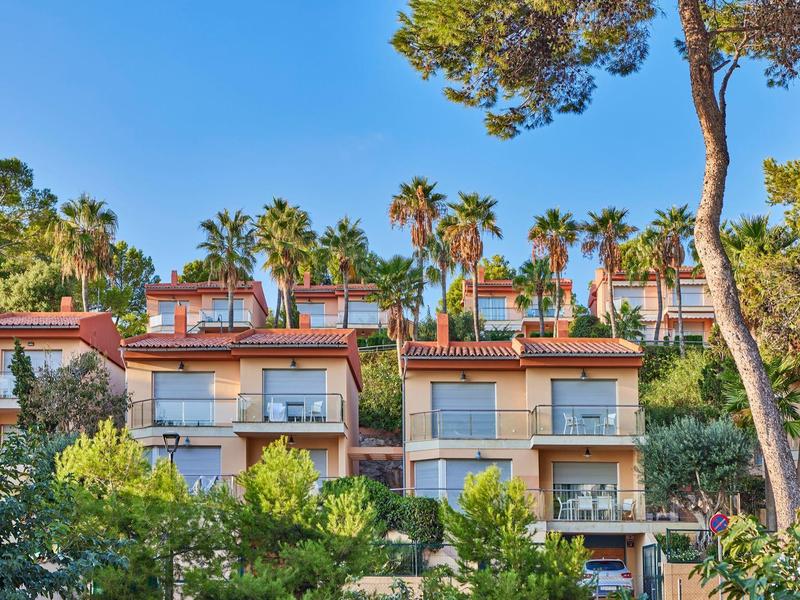 Meerdere moderne vakantiehuizen met balkons, omgeven door palmbomen en weelderig groen onder een blauwe lucht.
