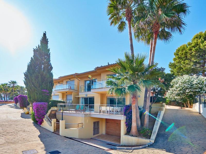 Twee verdiepingen tellend geel gebouw met balkon, palmbomen en bloeiende struiken langs een geplaveide weg.