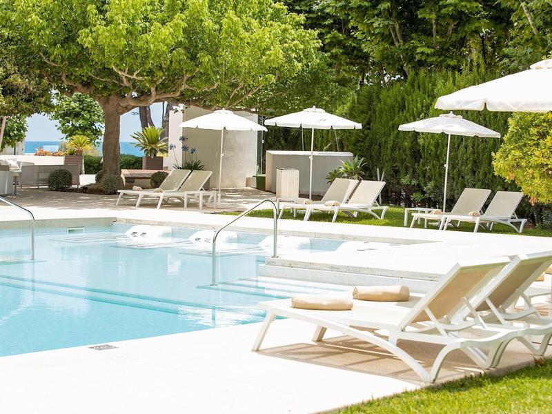 Schwimmbad mit weißen Liegestühlen, Sonnenschirmen und grünen Bäumen im Hintergrund.
