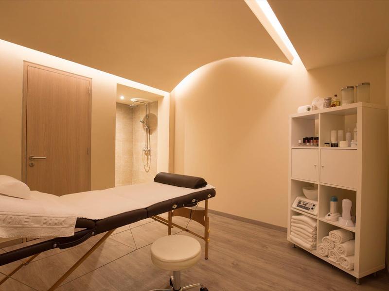 Helles Behandlungszimmer mit Massageliege, Hocker, Regalen und sanfter Deckenbeleuchtung.