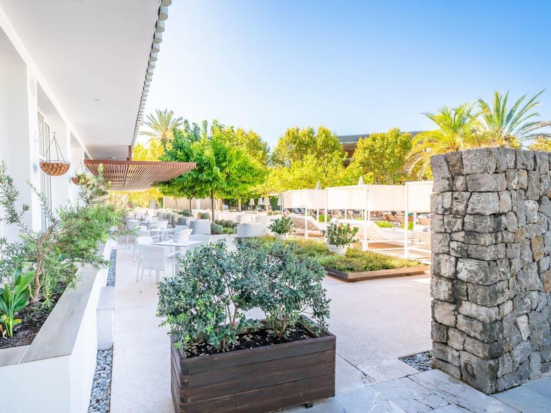 Moderner Garten mit weißem Boden, Steinmauer, Pflanzen in Kisten und Sonnenmarkise bei klarem Himmel.