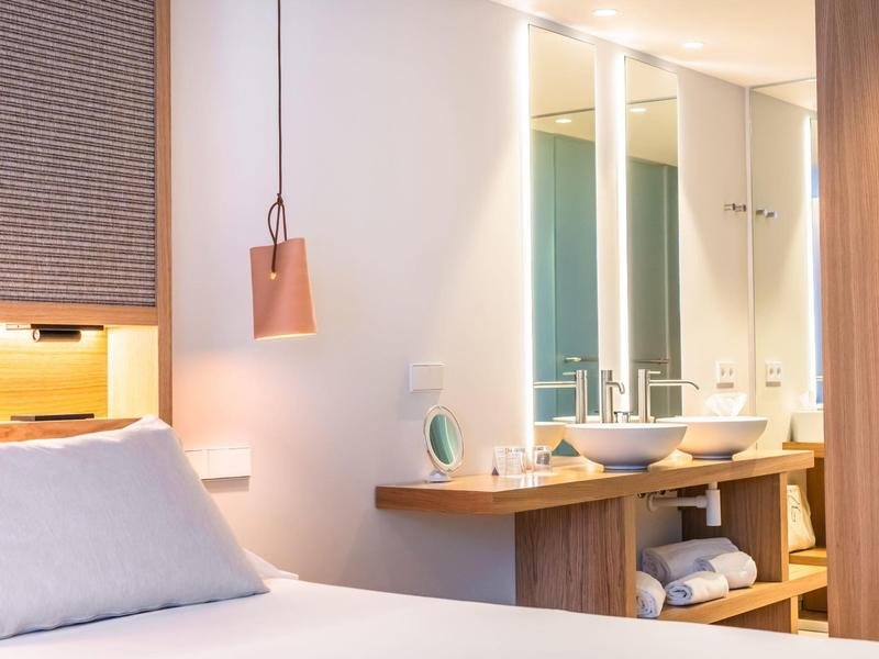 Helles Schlafzimmer mit weißem Bett, Holzmöbeln und modernem Waschbeckenbereich mit Spiegel.