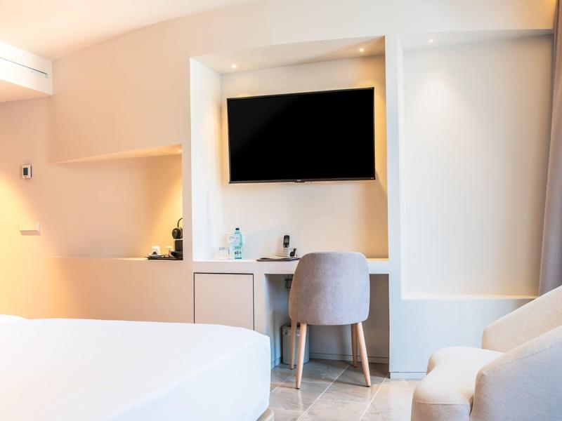 Helles, modernes Schlafzimmer mit Bett, grauem Stuhl, TV an der Wand und integriertem Schreibtisch.