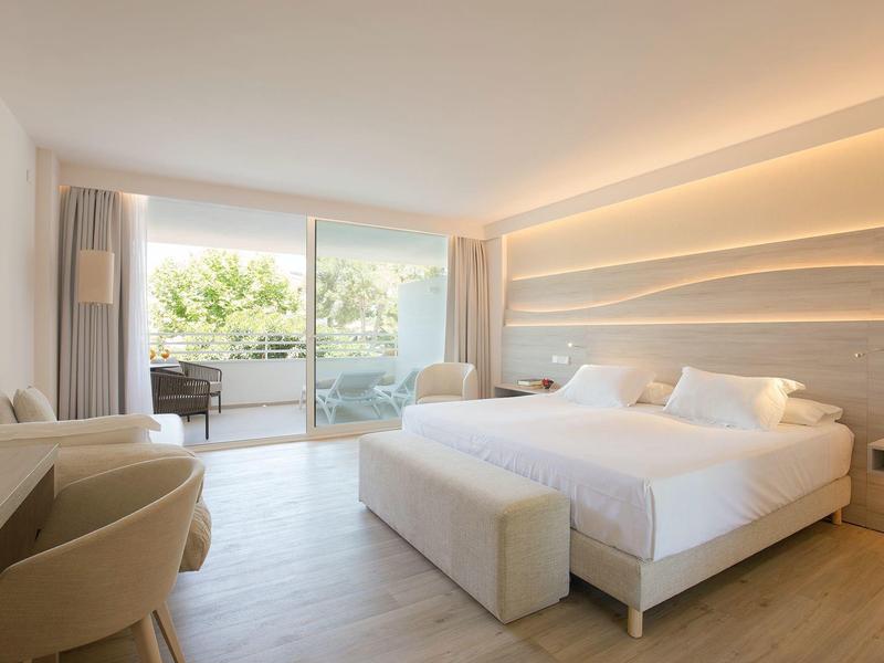 Helles Hotelzimmer mit Doppelbett, Sitzbereich, großen Fenstern und Balkon mit Aussicht auf Bäume.