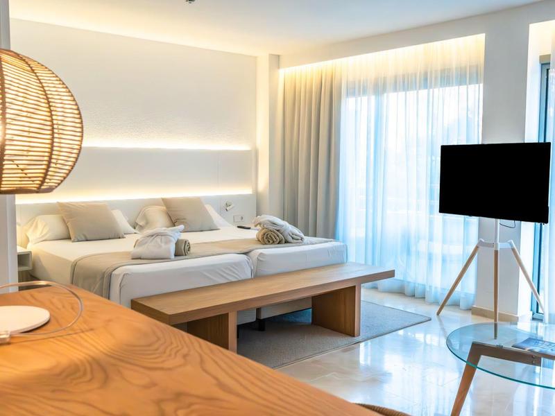 Helles Wohnzimmer mit weißem Sofa, großem Fenster, TV auf Stativ und runder Lampe auf Holztisch.