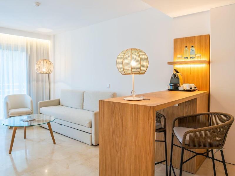 Heller Raum mit Holztheke, zwei Barhockern, Sofa, Sessel und großer Lampe am Fenster.