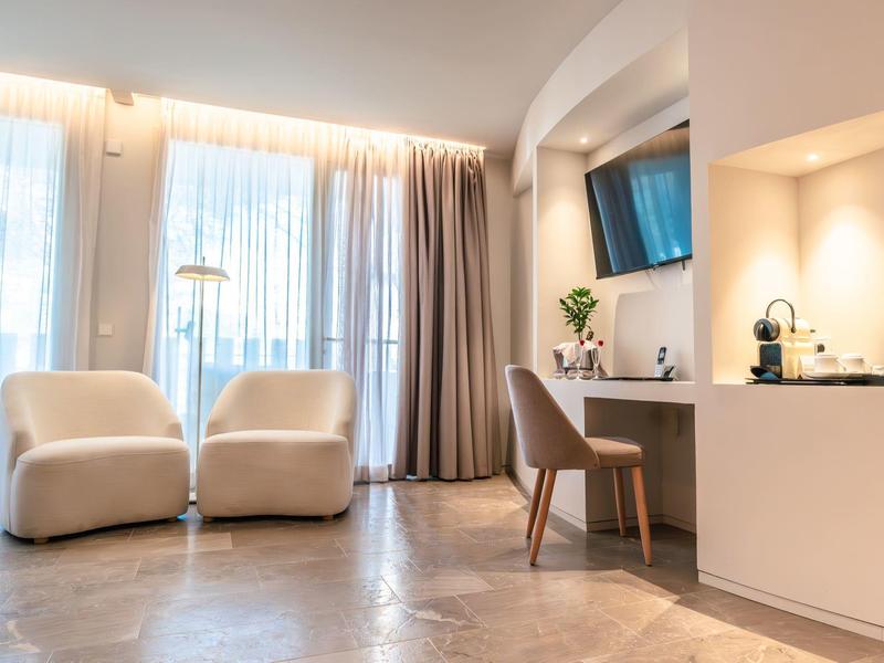 Helles Zimmer mit zwei Sesseln, Vorhängen, Schreibtisch, Stuhl, Fernseher und moderner Beleuchtung.