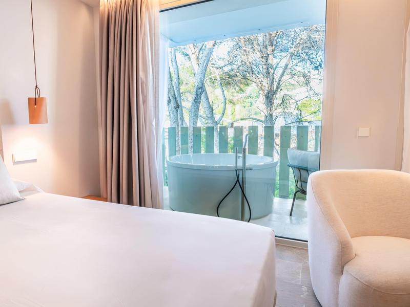 Helle Schlafzimmer mit großem Bett, beigem Sessel, Gardinen und Glasschiebetür zum Balkon mit Stuhl.