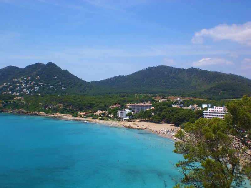 Klares blaues Meer vor grünen Hügeln und einem kleinen Küstenort unter blauem Himmel.