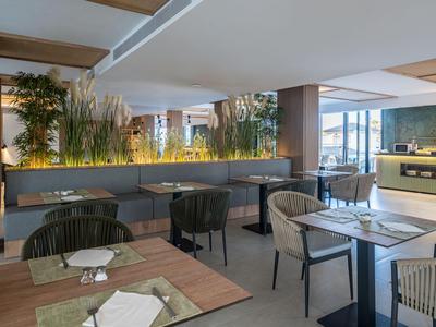 Ristorante d'hotel moderno con tavoli in legno chiaro, sedie e accento di piante verdi al centro.