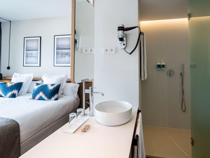 Moderní hotelový pokoj s manželskou postelí a otevřenou koupelnou se sprchou se skleněnými dveřmi.