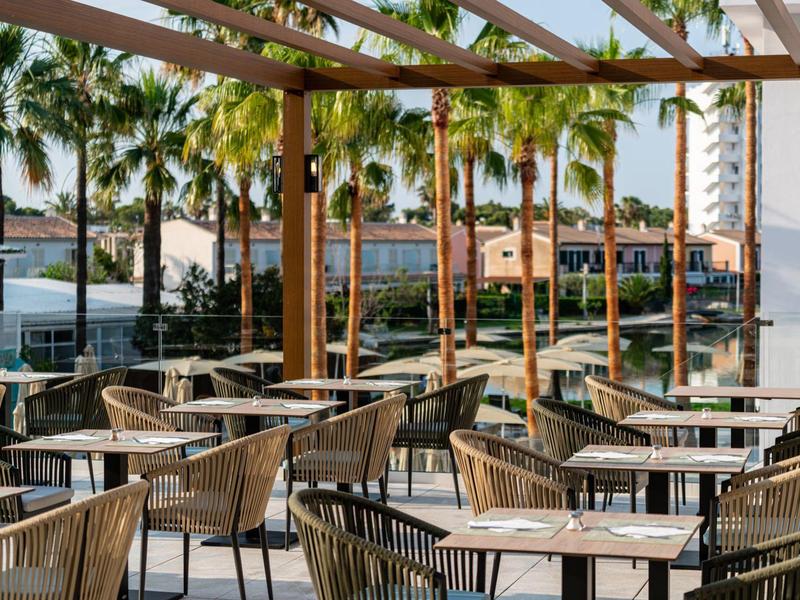 Terasa kavárny s dřevěnou pergolou, stoly, židlemi a palmami v pozadí.