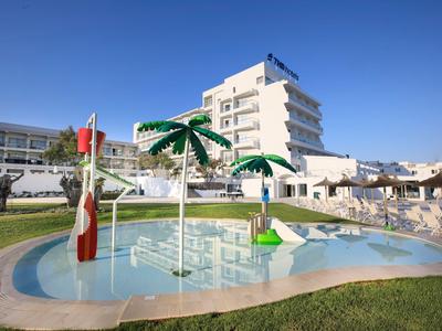 Piscina para niños con sombrillas y tumbonas frente a un edificio de hotel blanco bajo un cielo despejado.