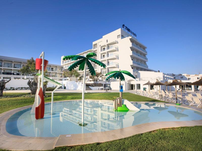 Piscine pour enfants avec palmiers, hôtel blanc en arrière-plan et ciel bleu clair.