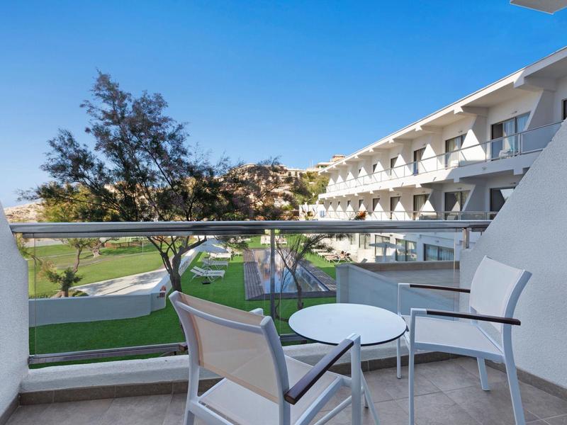 Balcon avec table ronde et chaises, vue sur jardin et bâtiment d'hotel sous un ciel bleu clair.