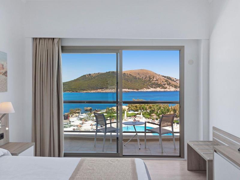 Chambre d'hôtel lumineuse avec balcon surplombant la mer turquoise et les collines.