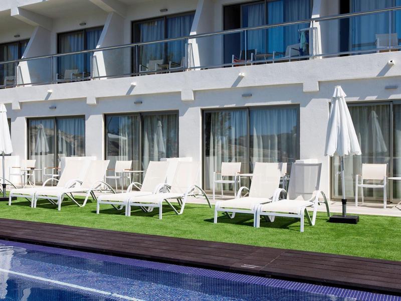 Chaises longues blanches près d'une piscine devant un hôtel moderne avec balcons et parasols.