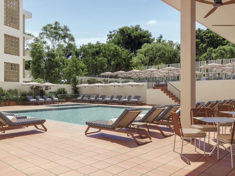 Hotel pool with loungers and tables under a shaded terrace on a sunny day