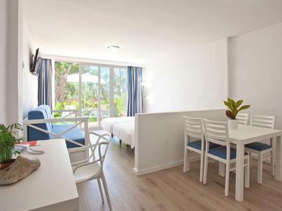 Camera d'hotel luminosa con letto, sedie blu, tavolo da pranzo, piante e vista sul balcone.