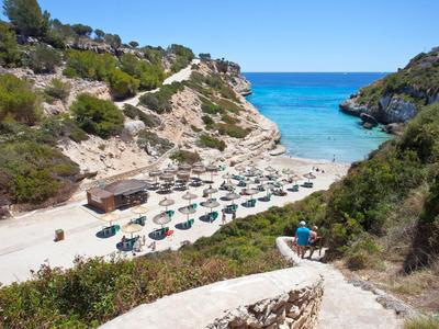 Spiaggia soleggiata con ombrelloni, acqua blu cristallina e colline verdi circostanti.