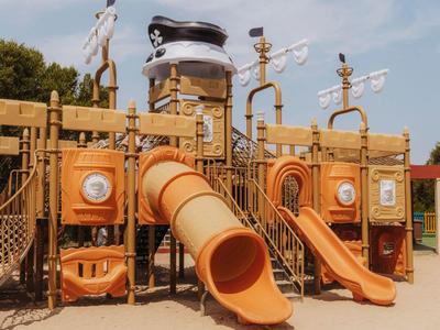 Gran parque infantil de madera con toboganes naranjas y diseño náutico en un día soleado.