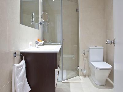 Bagno moderno compatto con doccia in vetro, WC bianco, mobile scuro e piastrelle beige.