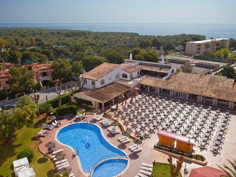 Un grande resort con piscina esterna curva, molte sdraio, circondato da verde e edifici.