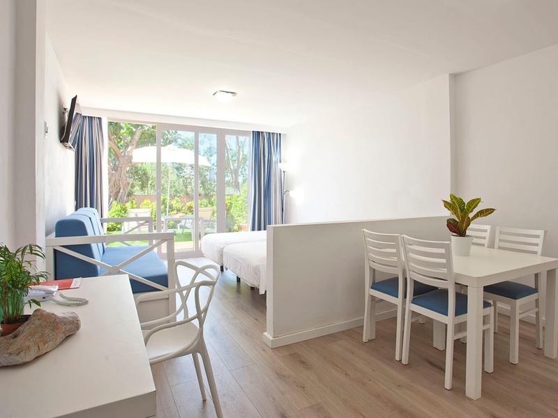 Camera d'hotel luminosa con letto, sedie blu, tavolo da pranzo, piante e vista sul balcone.