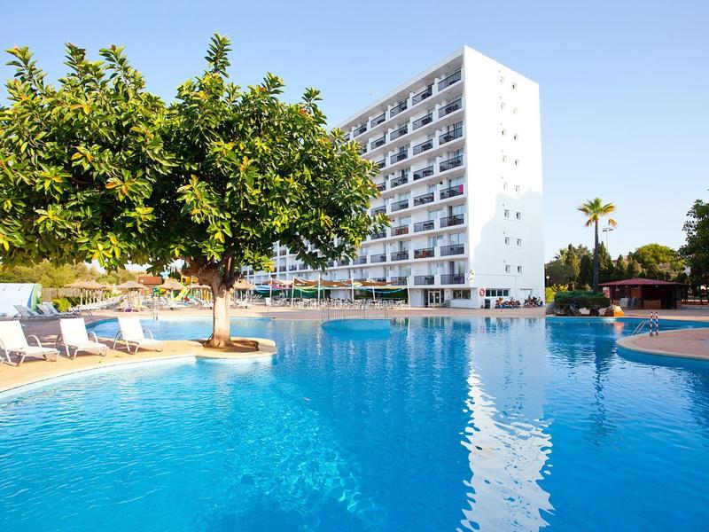 Hotel con una grande piscina all'aperto, lettini, un albero nell'acqua e cielo azzurro.