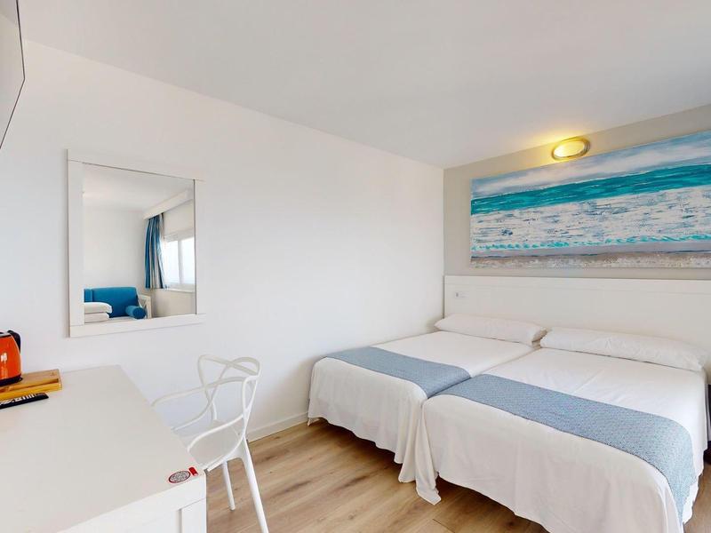 Camera d'hotel luminosa con due letti singoli, pavimento in legno, scrivania, sedia e decorazione murale a tema mare.