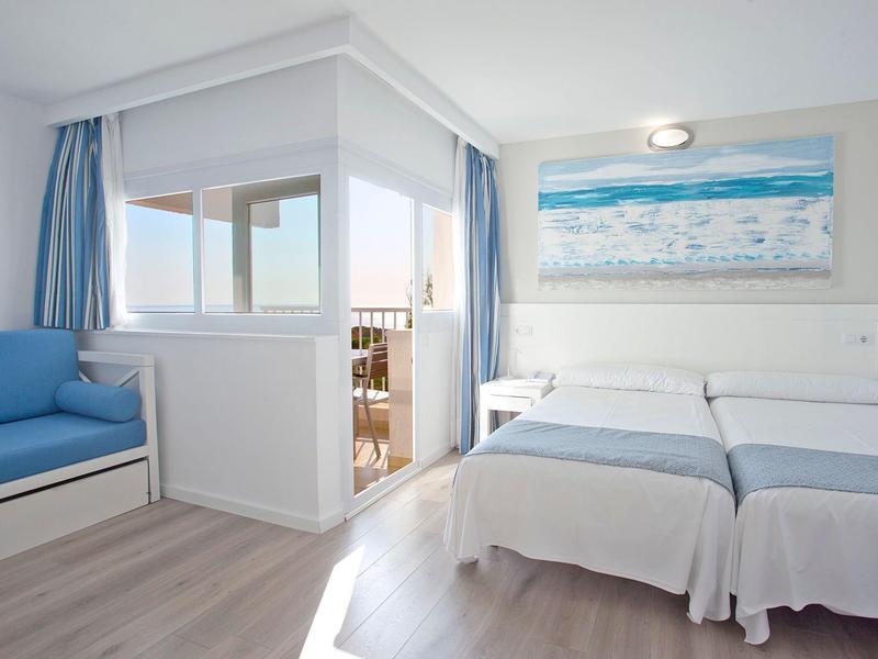 Camera d'hotel luminosa con due letti singoli, divano blu, balcone e quadro a tema mare.