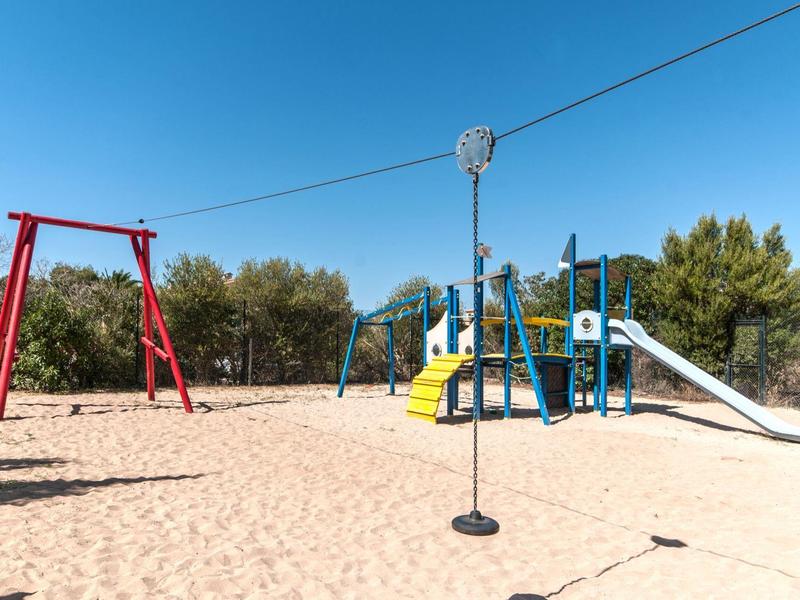 Area giochi soleggiata con altalene, scivoli e strutture per arrampicarsi sulla sabbia sotto un cielo azzurro.