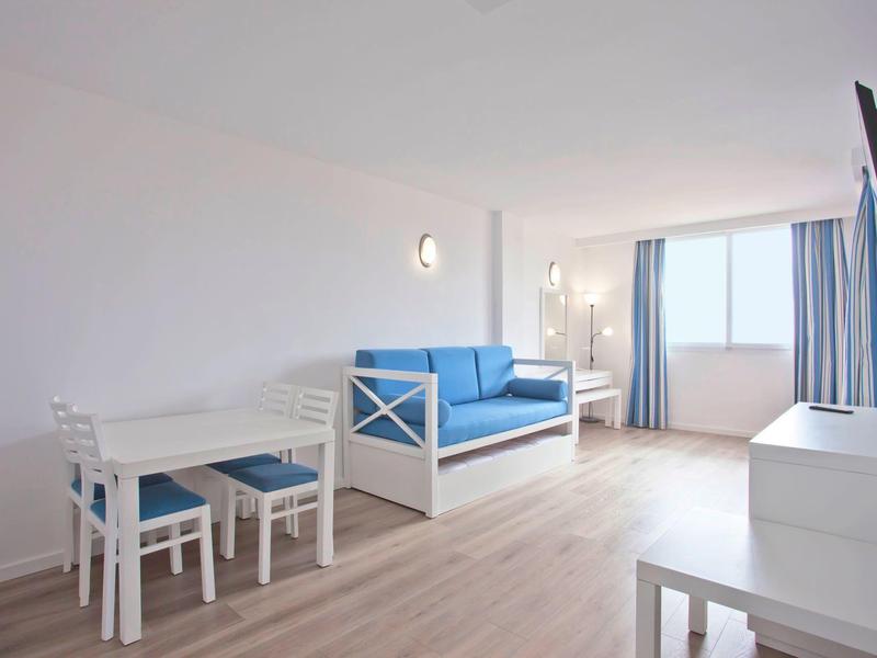 Camera d'hotel luminosa con divano blu, tavolo da pranzo bianco e grande finestra con tende.