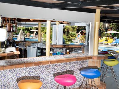 Bunte Barhocker vor offener Bar mit Fliesenmuster, Terrasse mit Liegestühlen und Sonnenschirmen.