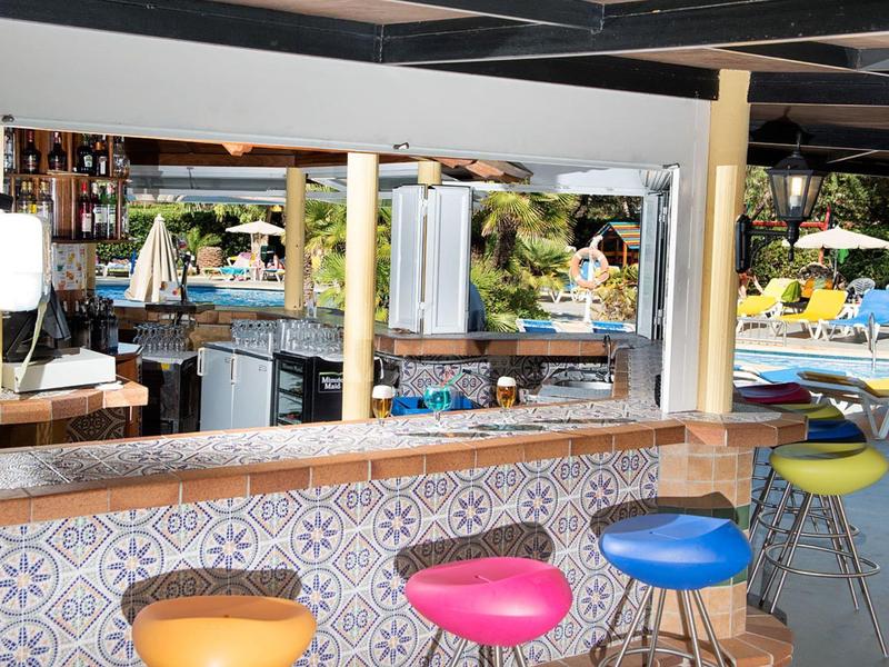 Bunte Barhocker vor offener Bar mit Fliesenmuster, Terrasse mit Liegestühlen und Sonnenschirmen.