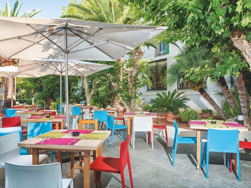 Bunte Stühle und Tische unter Sonnenschirmen in einem schattigen Außenbereich mit viel Grün.