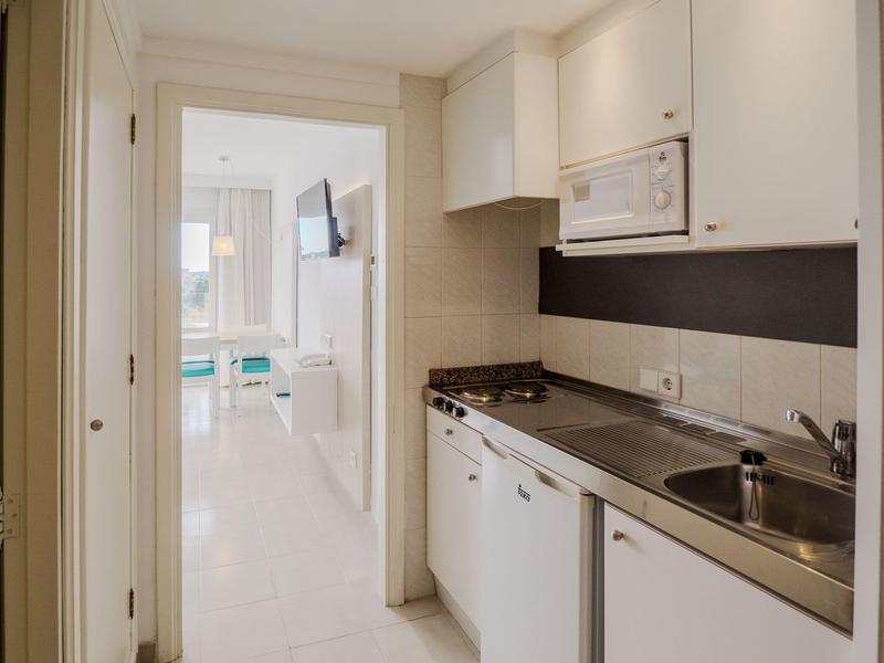 Kleine Hotelküche mit Herd, Spüle und Mikrowelle, Blick ins helle Wohnzimmer.